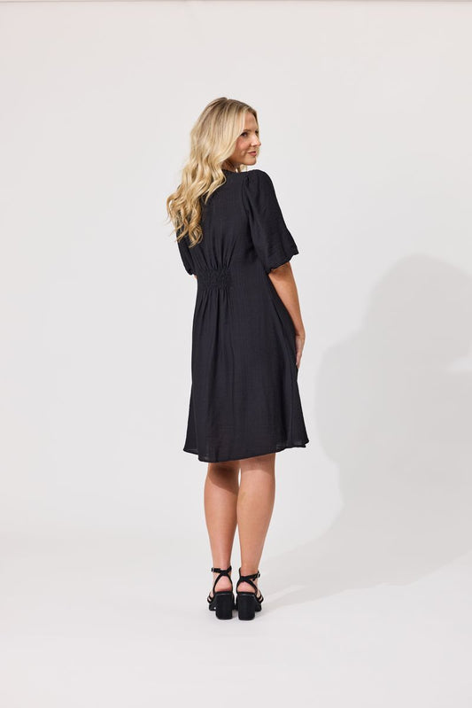 A woman modeling a black short-sleeve dress from the back, standing against a plain white studio background.