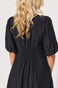 Back view of a woman wearing a black midi dress with puff sleeves and a smocked waist, shown against a plain light background.