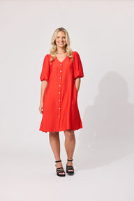 A woman modeling a bright red short-sleeve button-front dress with puff sleeves, paired with black strappy heels, against a plain light gray studio background.
