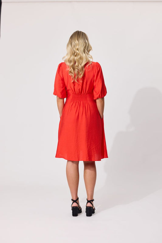 A woman viewed from behind wearing a bright red knee-length dress with short puff sleeves, a gathered waist, and black platform sandals against a plain white studio background.