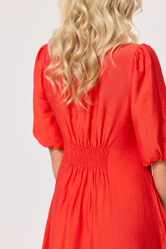 A woman wearing a bright red dress, shown from the back. The dress features short puff sleeves, a gathered smocked waist, and a flowing skirt.