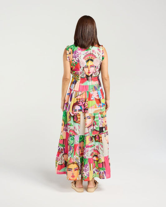A woman viewed from behind wearing a sleeveless, tiered maxi dress with a vibrant, multicolored collage print featuring illustrated faces. She is standing against a plain light gray studio background.