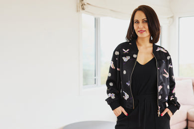 Woman wearing a black bomber jacket with butterfly patterns in a bright room.