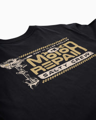 Close-up of a black crewneck t-shirt featuring a distressed gold and white graphic on the left chest that reads 'Motor Repair Salty Crew' with a wrench illustration.