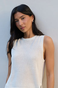 A woman with long dark hair wearing a ribbed white sleeveless knit top, posing against a light gray wall.