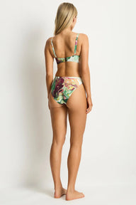 A woman stands barefoot against a plain light background, photographed from the back to show a two-piece bikini. The swimsuit has a colorful abstract print with green, pink, purple, and cream tones.
