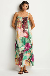 A woman modeling a sleeveless maxi dress with thin straps and a colorful abstract marble print in green, pink, purple, orange, and cream, standing against a plain light gray studio background.