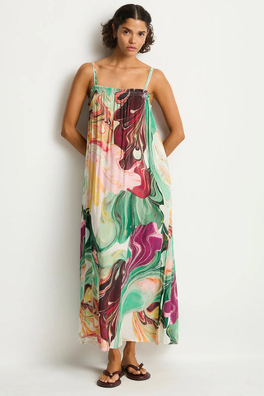 A woman modeling a sleeveless maxi dress with thin straps and a colorful abstract marble print in green, pink, purple, orange, and cream, standing against a plain light gray studio background.