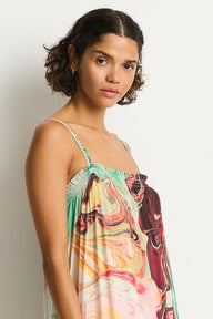 A woman wearing a sleeveless spaghetti-strap top with a colorful abstract swirl print in green, pink, orange, yellow, and burgundy, photographed against a plain light gray studio background.