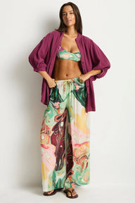 A woman modeling a matching two-piece swimwear and lounge set. The set features a green tropical print bikini top and matching wide-leg pants with a colorful abstract swirl pattern, styled with a draped magenta button-up shirt and brown sandals against a plain white background.
