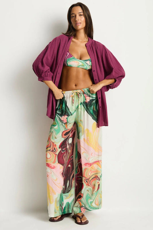 A woman modeling a matching two-piece swimwear and lounge set. The set features a green tropical print bikini top and matching wide-leg pants with a colorful abstract swirl pattern, styled with a draped magenta button-up shirt and brown sandals against a plain white background.