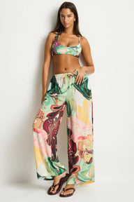 A woman modeling a matching two-piece swimsuit and wide-leg pants set with a colorful abstract swirl print in green, pink, yellow, and white, paired with dark brown flip-flops against a plain light gray studio background.