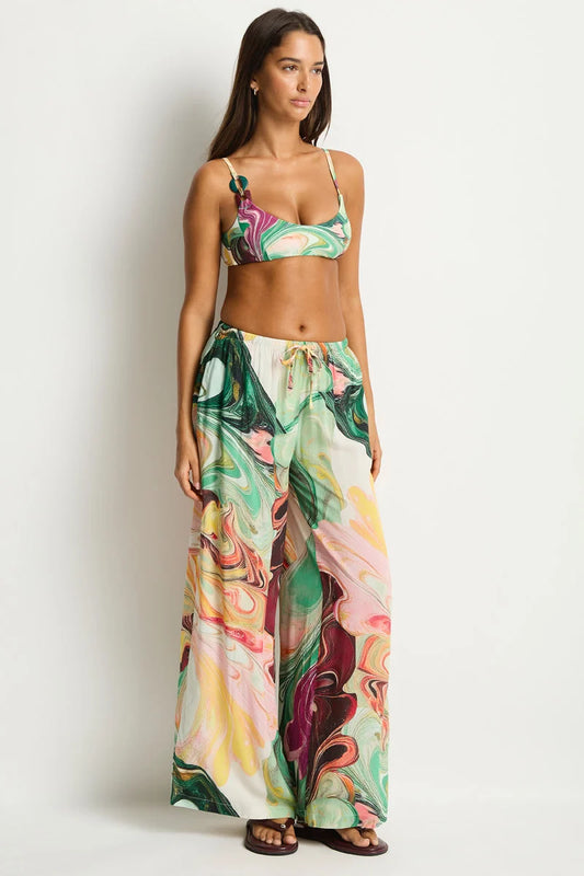 A woman modeling a matching two-piece swimwear set with a colorful abstract marbled print. The set includes a bikini top with thin straps and high-waisted, wide-leg maxi pants with a drawstring waist, photographed against a plain light gray studio background.