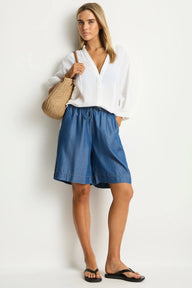 Woman wearing a white blouse and blue shorts holding a straw bag against a white background
