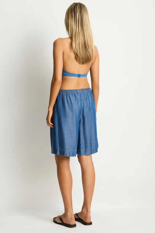 A woman shown from the back wearing a blue strapless bandeau top and matching blue shorts, standing against a plain light gray studio background.