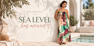 A woman wearing a colorful floral maxi dress stands in a sunlit outdoor patio setting next to a pool, with large text on the left reading 'NEW ARRIVALS SEA LEVEL has arrived'.