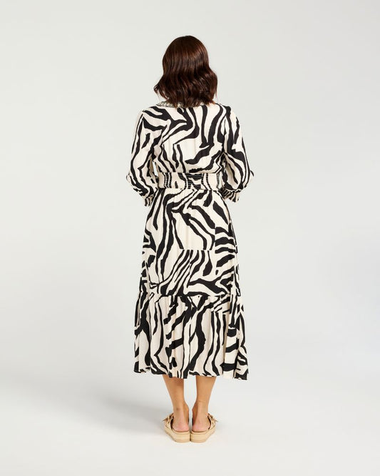 A woman viewed from behind wearing a long-sleeved midi dress with a bold black and white zebra print, paired with beige flat sandals, against a light gray studio background.