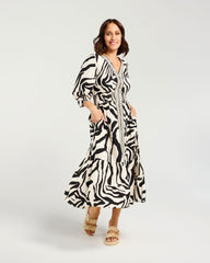 A woman modeling a black and white zebra-print midi dress with a belted waist, short puff sleeves, and a V-neckline, paired with beige strappy sandals against a light gray studio background.