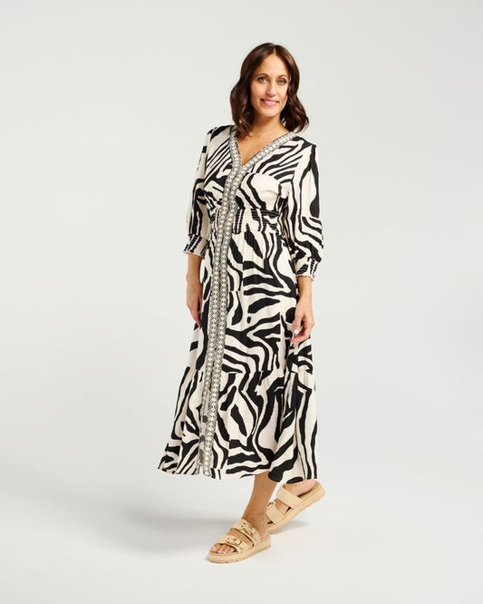 A woman modeling a black and white zebra-print midi dress with a silver beaded border down the front, paired with beige platform sandals, against a light gray studio background.