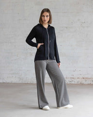 Woman wearing a black jacket and gray pants standing against a light gray brick wall.