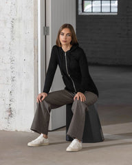 Woman sitting on a stool in a minimalistic indoor setting wearing a black hoodie and gray pants.