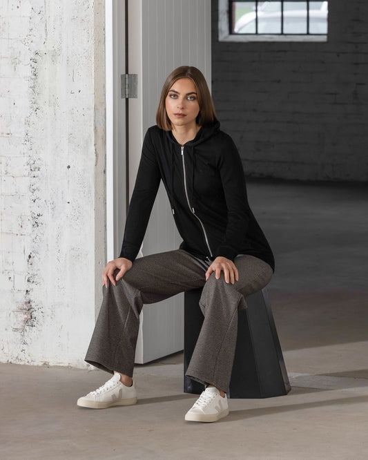 Woman sitting on a stool in a minimalistic indoor setting wearing a black hoodie and gray pants.