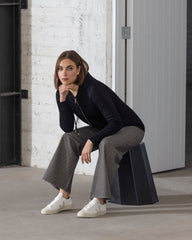Woman sitting on a black stool against a white brick wall.