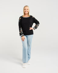 Woman wearing a black sweater with floral patterns and light blue jeans on a white background