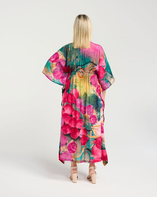 A woman viewed from behind wearing a long, loose kaftan-style dress with a vivid floral print in pink, teal, green, yellow, and blue, paired with beige strappy heels, against a light gray studio background.