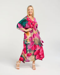 A woman modeling a long, flowing maxi dress with a vibrant abstract floral print in pink, green, and orange tones, paired with beige strappy high-heeled sandals against a light gray studio background.