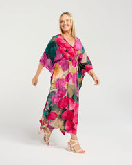 A woman modeling a long, flowy kaftan dress with a vibrant floral print in pink, green, and orange tones, paired with beige heeled sandals against a light gray studio background.