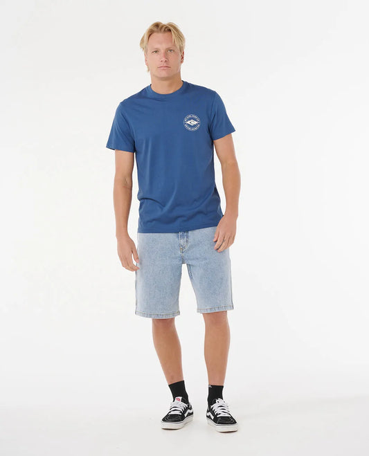 Man wearing a blue t-shirt with a logo and light blue shorts on a white background