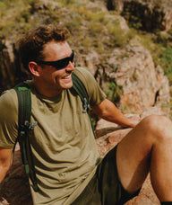 Man with sunglasses and a backpack sitting on a rock with a mountainous background