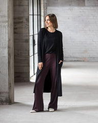 Woman wearing a black cardigan and dark purple pants standing in a room with concrete walls and a large window.