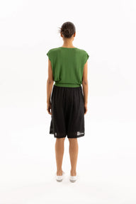 A model viewed from the back wearing a green sleeveless top and black shorts against a plain white studio background.