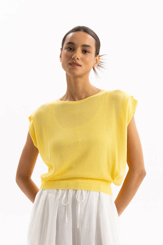 A woman wearing a bright yellow knit top with wide cap sleeves and a relaxed fit, paired with white drawstring trousers, photographed against a clean white studio background.