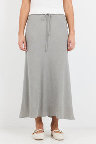 A model wearing a gray and white micro-patterned midi skirt with a drawstring waist, paired with a white top and white flat shoes, against a plain white background.