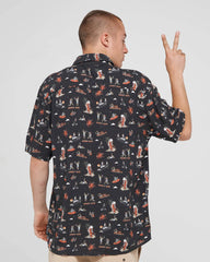 A man shown from the back wearing a black short-sleeve button-up shirt with an all-over graphic print of surfboards, people, and the words 'SUNSET BAY'. He is posing with one hand raised in a peace sign against a light gray studio background.