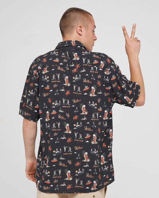 A man shown from the back wearing a black short-sleeve button-up shirt with an all-over graphic print of surfboards, people, and the words 'SUNSET BAY'. He is posing with one hand raised in a peace sign against a light gray studio background.