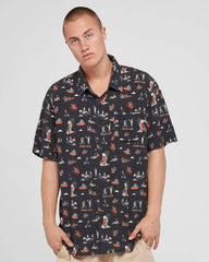 A man wearing a short-sleeve button-up shirt with a dark navy background and a colorful all-over retro illustration print, paired with beige pants, against a light gray studio background.