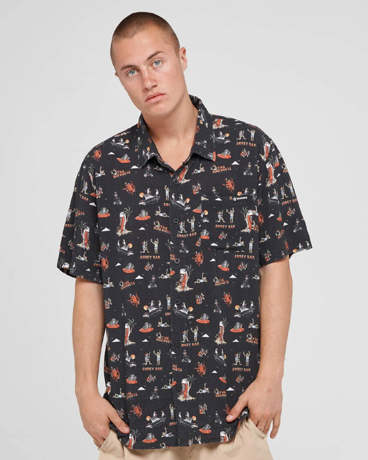 A man wearing a short-sleeve button-up shirt with a dark navy background and a colorful all-over retro illustration print, paired with beige pants, against a light gray studio background.