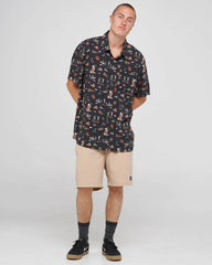 A man modeling a short-sleeve button-up shirt with a dark navy and orange fox pattern, paired with beige shorts, dark gray socks, and black sneakers, against a light gray studio background.