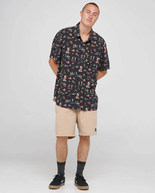 A man modeling a short-sleeve button-up shirt with a dark navy and orange fox pattern, paired with beige shorts, dark gray socks, and black sneakers, against a light gray studio background.