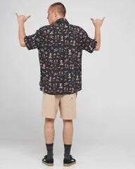 A man shown from the back wearing a short-sleeve button-up shirt with a multicolor cartoon dog pattern on a dark background, paired with beige shorts, black socks, and black shoes, posing with his hands raised against a light gray studio background.