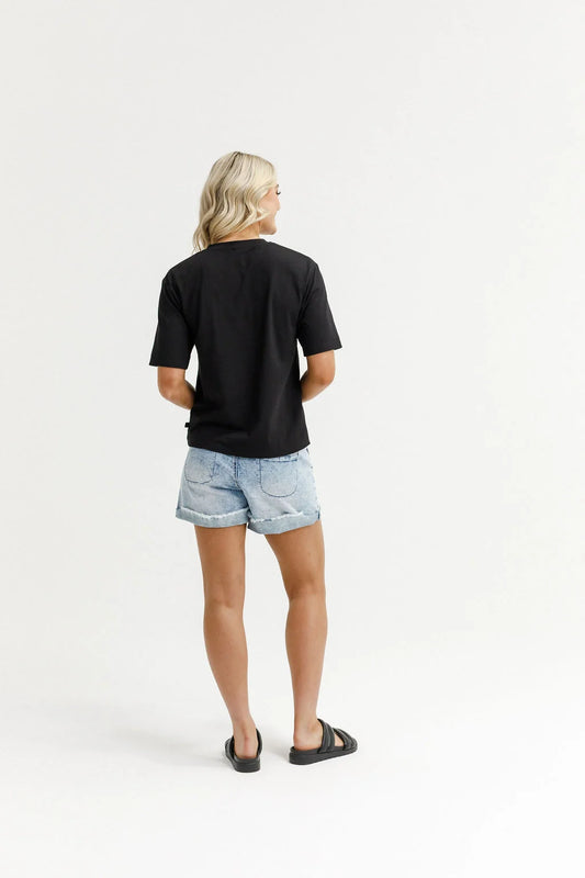A woman viewed from behind wearing a black short-sleeve t-shirt, light-wash denim shorts, and black slide sandals, standing against a plain white studio background.