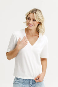 A smiling blonde woman wearing a white short-sleeve V-neck top and light blue jeans, posing against a clean white studio background.