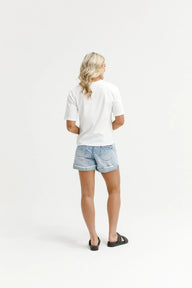 A woman shown from behind wearing a white short-sleeve t-shirt, light-wash denim shorts, and black slide sandals, standing against a seamless white studio background.