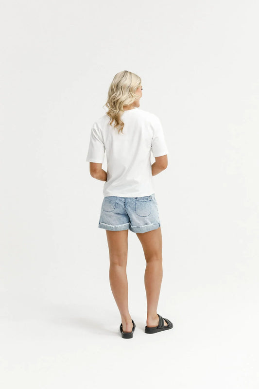 A woman shown from behind wearing a white short-sleeve t-shirt, light-wash denim shorts, and black slide sandals, standing against a seamless white studio background.