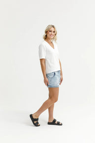 A woman modeling a white short-sleeve V-neck t-shirt and light-wash denim shorts, paired with black slide sandals, against a plain white studio background.
