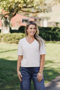 Lady wearing a white v neck tee & jeans standing in a garden under a big tree